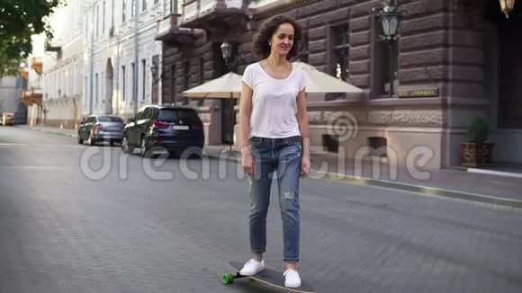 日出时穿着白色t恤蓝色牛仔裤和白色运动鞋的漂亮黑发女人在城市街道上玩滑板视频的预览图