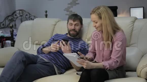 沙发上的金发妻子在笔记本上读笔记丈夫坐在旁边打电话视频的预览图