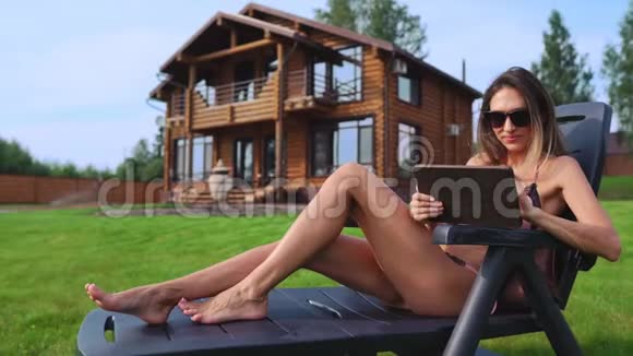 一个美丽苗条的女人躺在阳光下的休息室里穿着一件泳衣在一座大别墅的背景上放着一块平板电脑视频的预览图