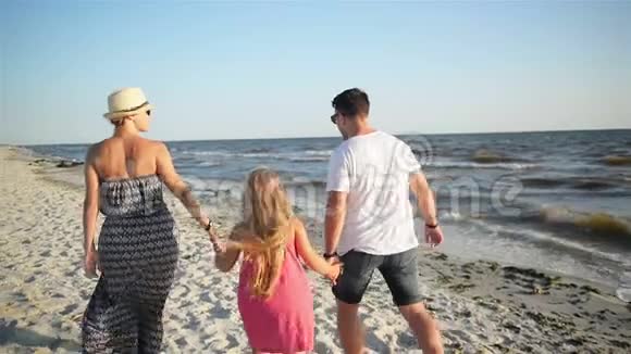 在夏日度假期间在沙滩上牵手散步快乐夫妻和小女儿的后景视频的预览图
