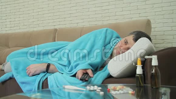 一个有感冒症状的成年男性躺在沙发上的毯子里用遥控器打开电视频道视频的预览图