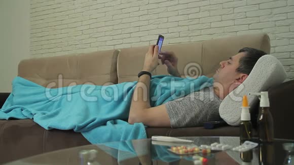 一个有冷躺迹象的成年男子裹在沙发上的毯子里手里拿着一部手机视频的预览图