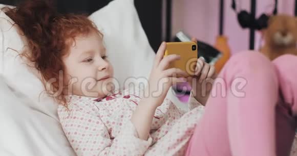 有趣的红发小女孩躺在床上饶有兴致地在现代黄色智能手机上观看视频视频的预览图