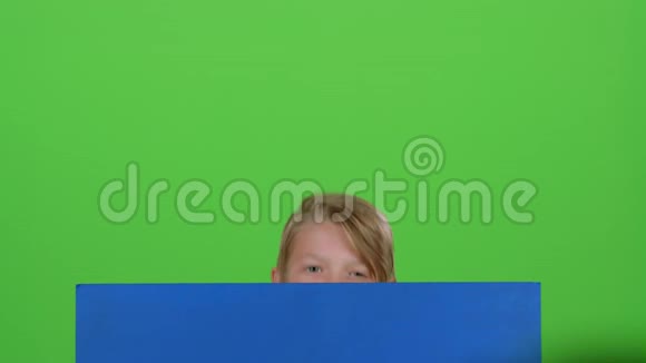 穿套头衫的小男孩从黑板后面出来在绿色的屏幕上挥手视频的预览图