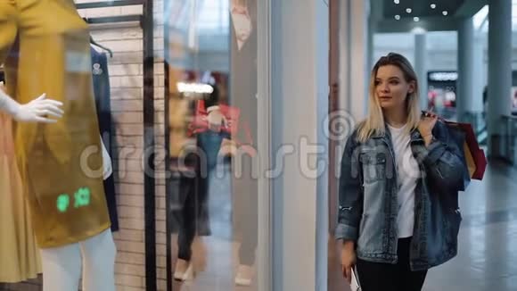 一个年轻的金发女郎带着袋子在商场里沿着橱窗走着视频的预览图