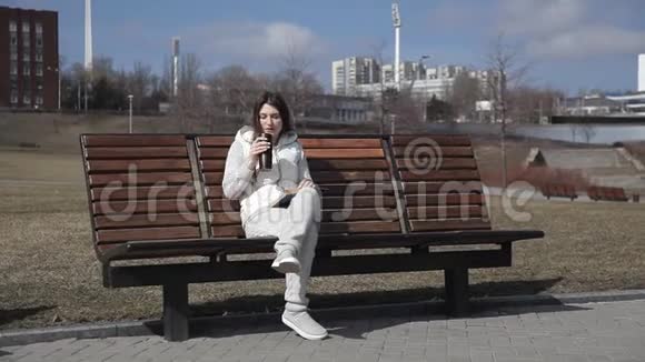 一个穿着运动服的漂亮女孩坐在长椅上看书喝着热杯里的咖啡视频的预览图