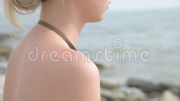 一个迷人的年轻女孩在阳光明媚的夏天坐在海滨在她的肩膀上涂上防晒霜夏季视频的预览图