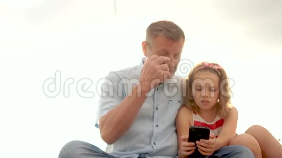 一个成熟的男性养老金领取者和一个小卷曲的女孩祖父和孙女在智能手机上玩游戏笑了起来视频的预览图