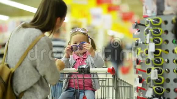 长着长发的布鲁内特女士为她的小女儿选择太阳镜站在货架附近两个可爱的女孩视频的预览图