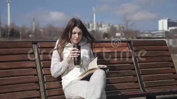 一个穿着运动服的漂亮女孩坐在长椅上看书喝着热杯里的咖啡视频的预览图