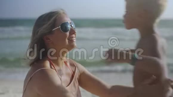 年轻的怀孕母亲和儿子坐在沙滩上在夏天亲吻和玩耍视频的预览图