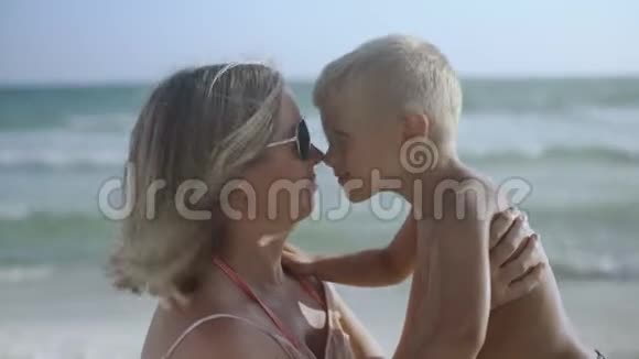 年轻的怀孕母亲和儿子坐在沙滩上在夏天亲吻和玩耍视频的预览图