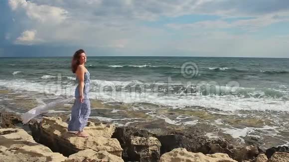 快乐的年轻女子在多风的天气里在岩石海滩上放松看着相机视频的预览图
