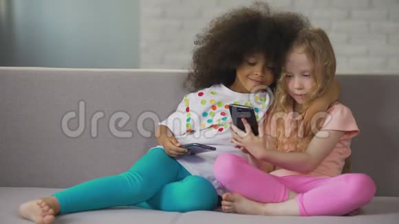 多种族姐妹坐在沙发上拥抱并在智能手机上自拍视频的预览图