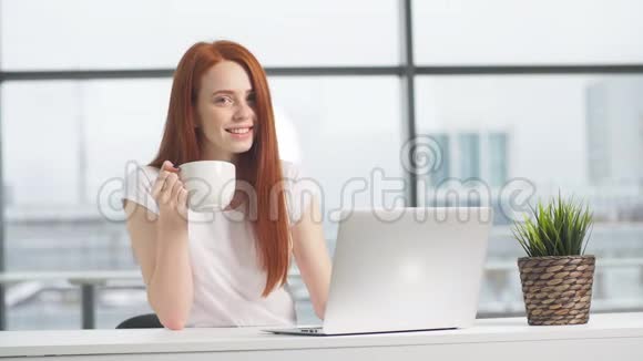 快乐的红头发女孩坐在桌子旁手提电脑看着相机视频的预览图