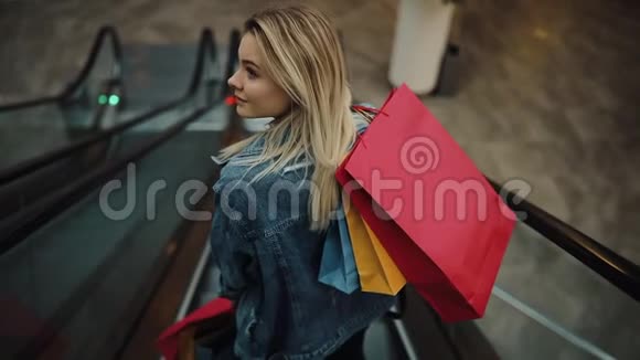 穿着牛仔裤夹克的年轻金发女人在购物中心的移动楼梯上倒下视频的预览图