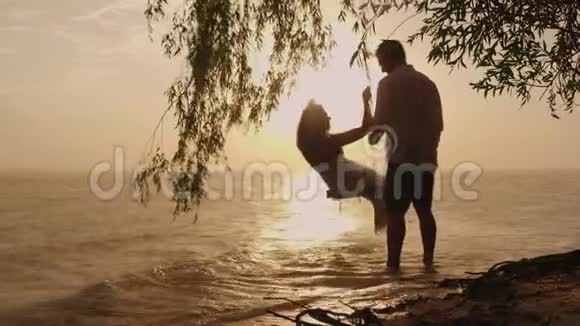 一对年轻夫妇的剪影一个男人在秋千上滚动他的妻子在日落时大海的背景下它了视频的预览图
