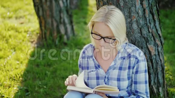 一个戴眼镜的金发年轻女子在公园里看书位于一棵树附近日落前美丽的灯光顶部视图视频的预览图