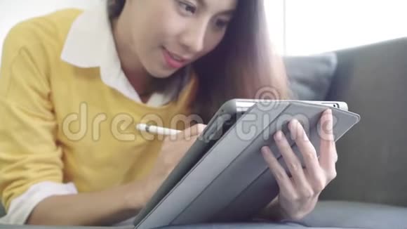 美丽的亚洲女性使用平板电脑观看视频而穿毛衣躺在沙发上的客厅在家里家庭生活方式妇女视频的预览图