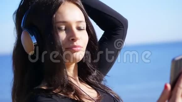 夏天在大海的背景下美丽的女人留着长长的黑发听着耳机里的音乐视频的预览图