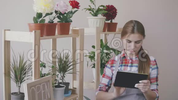 小生意一个穿着条纹围裙和格子衬衫的女孩站在一家花店里架子上有花和一朵视频的预览图