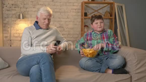 爷爷和孙子坐在沙发上看电视吃薯片一个年长的男人视频的预览图