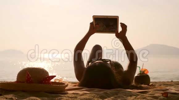 年轻的旅游女孩躺着在热带海滩晒日光浴使用移动平板小玩意4K慢动作泰国普吉岛视频的预览图