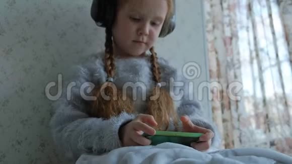 快乐的小女孩戴着耳机的辫子用电话玩微笑坐在房间的窗台上视频的预览图