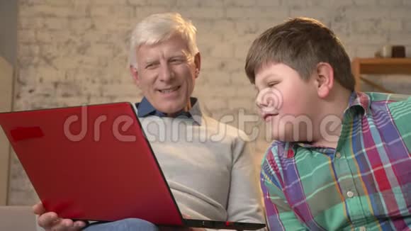 祖父和孙子坐在沙发上在笔记本电脑上看着一部有趣的电影笑着说着家庭视频的预览图