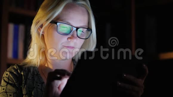 中年妇女工作到很晚享受平板电脑平板电脑的屏幕反映在她的眼镜上视频的预览图