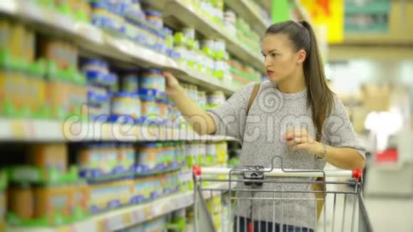 令人惊叹的布鲁内特母亲与购物车购买健康婴儿食品在超市站在货架附近视频的预览图