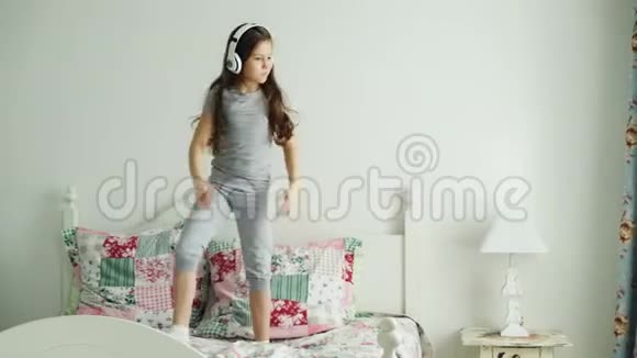 有趣可爱的小女孩戴着无线耳机跳舞在假期中享受乐趣早上在舒适的家里跳床视频的预览图