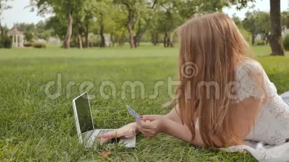 一个年轻的学生女孩躺在城市公园的草地上用信用卡和笔记本电脑付款视频的预览图