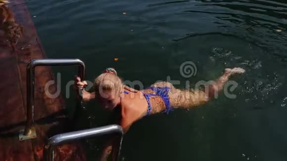 女人戴着太阳镜和泳衣坐在码头上女孩在洪水木水下码头休息在水里的女士视频的预览图