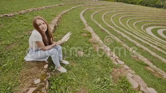 一个年轻的女学生正在看一本书坐在一个大体育场的草地上准备学习视频的预览图