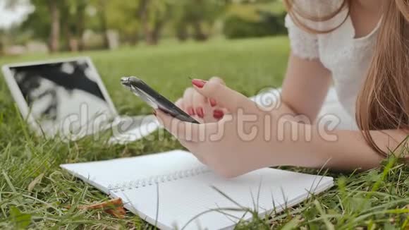 一个年轻的学生女孩躺在公园的草地上用便携式电脑做笔记学习筹备工作视频的预览图