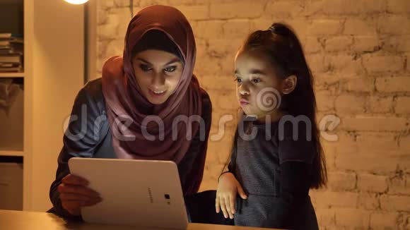 晚上一位戴着头巾的年轻母亲和她的小女儿坐在沙发上用平板电脑看着一个有趣的东西视频的预览图