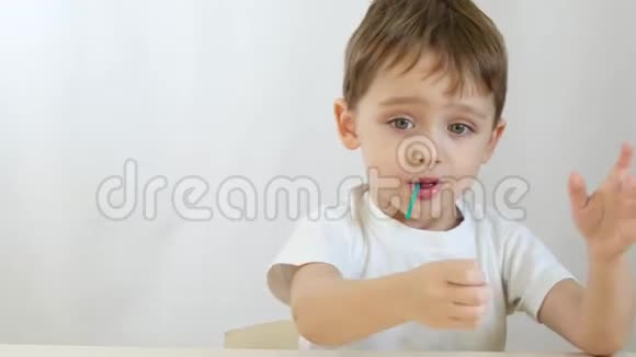 快乐的小男孩微笑着在棍子上舔棒棒糖坐在白色背景的桌子旁视频的预览图