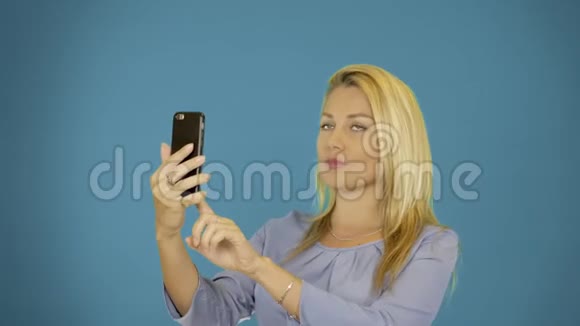年轻的白种女人在智能手机上拍照纯蓝色背景女性肖像齐腰漂亮的金发女郎微笑着视频的预览图