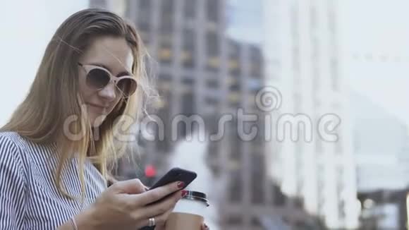 年轻女商人戴太阳镜使用智能手机的肖像休息时拿着咖啡杯在街上视频的预览图