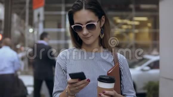 一幅年轻漂亮的女商人的肖像她拿着咖啡杯在拥挤的街道上用智能手机记录视频的预览图