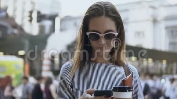 年轻漂亮的女商人站在拥挤的街道上拿着咖啡和文件用智能手机视频的预览图