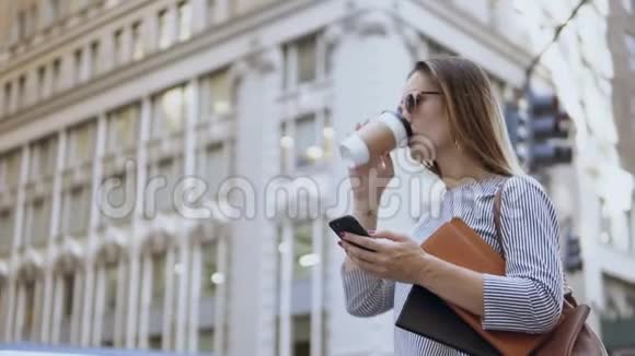 年轻忙碌的女商人戴着太阳镜拿着文件在街上使用智能手机和喝咖啡视频的预览图