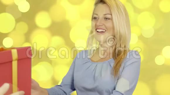 快乐的白种人女人在黄金背景下拿红色礼品盒微笑的女人带来惊喜视频的预览图