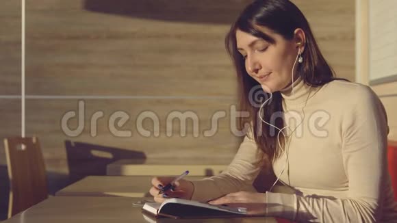 年轻女子拿着耳机坐在咖啡馆里在笔记本上做笔记视频的预览图