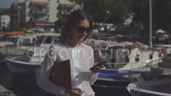 商务女性穿着正式服装的智能手机站在码头上背景是模糊的船只并打印出一张照片视频的预览图