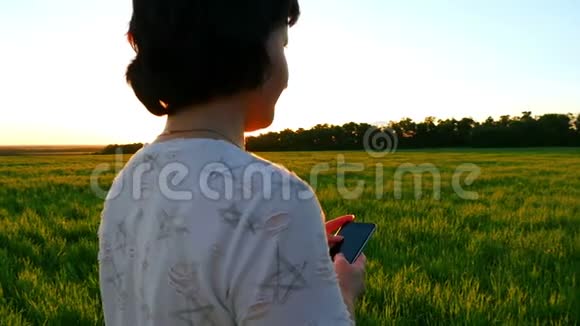 一位年轻的女士正拿着一部智能手机在夕阳的背景下在一片绿色的草坪上慢动作着视频的预览图