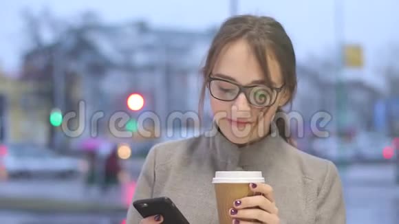 微笑的年轻女性在喝咖啡时检查智能手机上的社交媒体视频的预览图