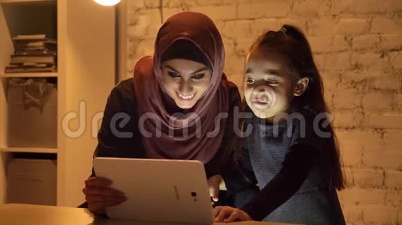 晚上一位戴着头巾的年轻母亲和她的小女儿坐在沙发上用平板电脑看着一个有趣的东西视频的预览图