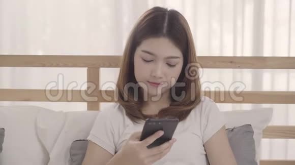 年轻的亚洲女人在早上醒来后躺在床上使用智能手机视频的预览图